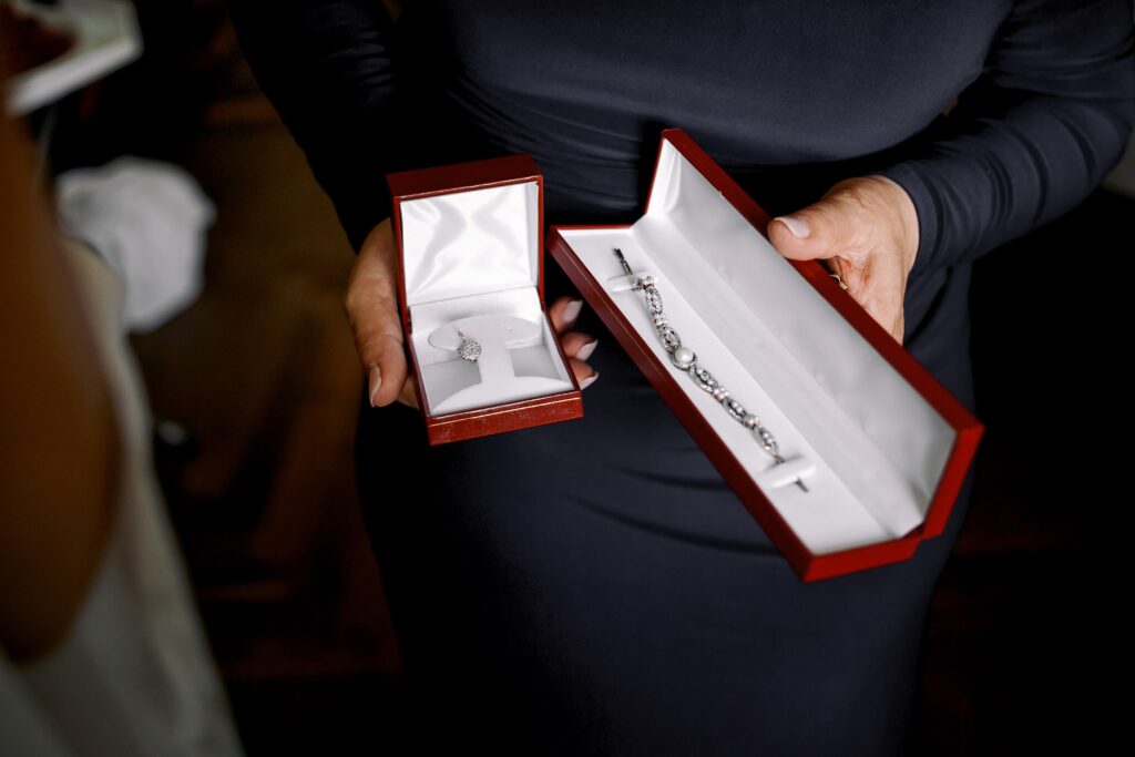 Kvinna i mörk klänning håller i två smyckesaskar i röd sammet – en med ett örhänge i silver och en med ett diamantarmband.